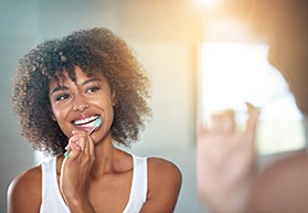 Woman in white tank top at bathroom mirror brushing teeth
