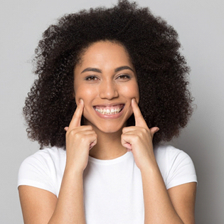 A young woman wearing a white shirt and using her index fingers to point to her cheeks while she smiles