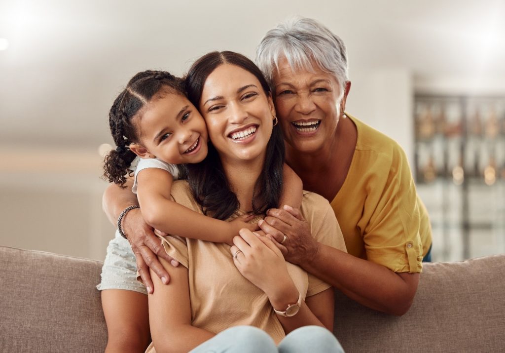 Grandmother, mom, and daughter smiling while hugging on couch